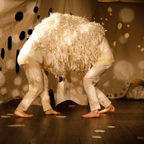 grafika przedstawia dwie bose, pochylone postaci stojące przodem do siebie, na głowach i barkach mają narzuconą narzutę z długimi frędzlami