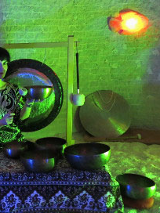 Kobieta trzyma w ręku misę i w nią uderza. Na niskim stoliczku przykrytym suknem misy. Z tyłu wisi gong i oparty gong przy ścianie.