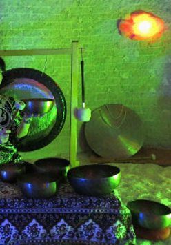 Kobieta trzyma w ręku misę i w nią uderza. Na niskim stoliczku przykrytym suknem misy. Z tyłu wisi gong i oparty gong przy ścianie.