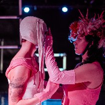 Na ciemnym tle dwie postaci w różowych strojach, stoją bokiem do widza, przodem do siebie.