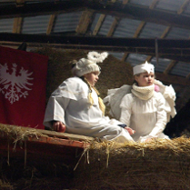 Dwie dziewczynki w strojach aniołów siedzą na belce w stodole, za nimi widać zawieszoną na ścianie czerwoną flagę z białym orłem.