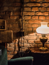 Stara miedziana lampa z ozdobnym kloszem stoi na stoliku. Oświetla zielony fotel, rozwieszoną gazetę i stare radio. W tle ceglana ściana.