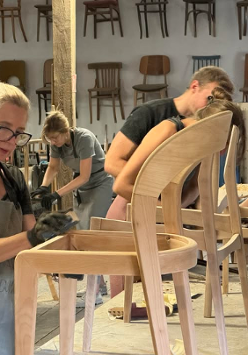 Na zdjęciu widać grupę osób pracujących nad drewnianymi krzesłami w warsztacie. Na obrazie znajduje się cztery osoby, wszystkie ubrane w codzienne ubrania i pracujące nad różnymi aspektami krzeseł. Krzesła są wykonane z jasnego drewna i mają prosty, nowoczesny design. W warsztacie rozrzucone są narzędzia i materiały, a ściany ozdobione są półkami z gotowymi krzesłami. Osoby na zdjęciu są skoncentrowane na swojej pracy, a atmosfera jest pełna twórczości i rzemiosła.