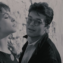 Czarno-biały kadr kobiety w słońcu opierającej się o ścianę, a obok niej mężczyzna w okularach.