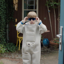 Chłopiec w stroju kosmonauty dotyka założonych przez siebie ciemnych okularów. Stoi na podwórku, w tle różne przedmioty oraz pnący się po ścianie drewnianego budynku bluszcz. Na pierwszym planie, po prawej stronie otwarte czerwono-niebieskie drzwi.