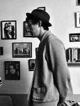 Czarno-białe zdjęcie mężczyzny i kobiety stojących przodem do siebie i rozmawijających w salonie. Za nimi ściana z fotografiami i kanapa.