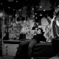 Mężczyzna leżący na łózku. Nad łóżkiem ściana pełna plakatów. przed nimi dziewczyna wybierająca płytę przy gramofonie. Zdjęcie jest czarno-białe.