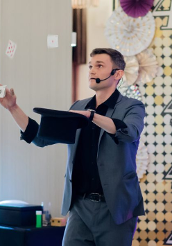 na zdjęciu mężczyzna w ciemnym garniturze, w jednej ręce trzyma czarny kapelusz, w drugiej talię kart