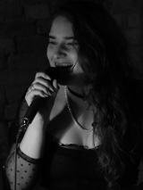 Zdjęcie, dziewczyna z mikrofonem w ciemnym pomieszczeniu. Dziewczyna ma ropuszczone długie włosy, koronkową bluzkę i naszyjnik z pereł.