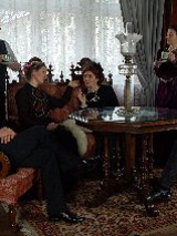 Zdjęcie, kadr filmu, aktorzy siedzą na eleganckich fotelach w salonie, w dłoniach trzymają filiżanki, przed nimi drewniany rzeźbiony stół.