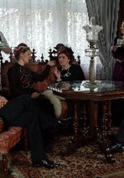 Zdjęcie, kadr filmu, aktorzy siedzą na eleganckich fotelach w salonie, w dłoniach trzymają filiżanki, przed nimi drewniany rzeźbiony stół.