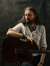 Mężczyzna w średnim wieku z długimi ciemnymi włosami i brodą. Ubrany w białą luźną koszulkę. Siedzi na wysokim krześle i jedną rękę opiera na gitarze akustycznej. Na ręce i na szyi ma biżuterie. Spogląda w bok.