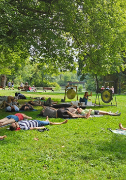 Zdjęcie, polana w parku, wokół drzewa, ludzie leżą na karimatach, przed nimi artyska gra na gongach i misach.