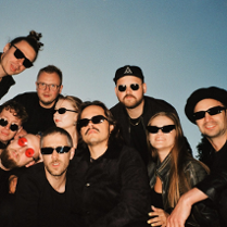 Zdjęcie grupy artystów stojących na tle nieba. Część osób kuca, część jest pochylona. Większość ma ciemne okulary. Wszyscy ubrani są w czarne ubrania.