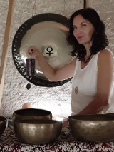 Zdjęcie przedstawia kobietę siedzącą przed dużym gongiem oraz kilkoma misami do śpiewu. Kobieta trzyma mały, czarny, prostokątny przedmiot w swojej prawej ręce. Ma na sobie białą koszulkę bez rękawów i naszyjnik. Gong jest zawieszony na suficie za kobietą. Misy do śpiewu są ułożone w półkolu przed nią. Tło stanowi biała ściana. Kobieta ma ciemne włosy i patrzy w obiektyw aparatu. Zdjęcie wydaje się być wykonane w pomieszczeniu z białą ceglaną ścianą. Kobieta lekko się uśmiecha.