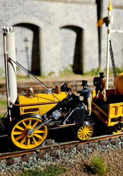 To jest miniaturowy model parowozu Rocket, historycznego pociągu, który zrewolucjonizował transport. Został ustawiony na makiecie torów kolejowych, kompletny z miniaturowym wagonem węglowym i figurkami ludzi. Model jest bardzo szczegółowy, z dokładnymi reprezentacjami projektu lokomotywy, w tym kotła, kół i nawet kominu. Model jest również pomalowany w jaskrawych kolorach, co dodaje realizmu. Tło zdjęcia wydaje się być miniaturowym modelem kamiennego muru, z arkadami w tle, dodającym do poczucia skali.