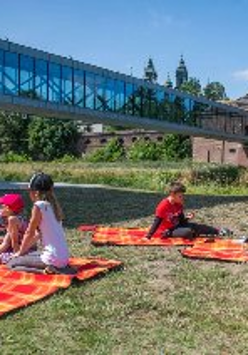 Grupa dzieci leży na kocach na trawniku. W tle widać szklaną kładkę prowadzącą nad rzeką do starego, ceglanego budynku.