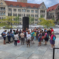 Zdjęcie, grupa osób stoi przed pomnikiem, za nimi ulica, drzewa, dwa samochody.