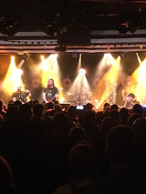 Na pierwszym planie głowy widowni, która słucha koncertu. Na oświetlonej scenie piątka mężczyzn w średnim wieku gra koncert na różnych instrumentach. Wśród nich jest gitarzysta, bębniarz, wiolonczelista, akordeonista.