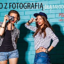 Przed niebieskim murem stoją w stylizowanych pozach dwie dziewczyny. Jedna trzyma w dłoniach aparat. Na ścianie napisy, kóre informują o wydarzeniu.