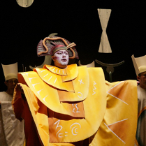 Śpiewak operowy stoi na scenie ubrany w żółty kostium. W tle kilku innych uczestników opery.