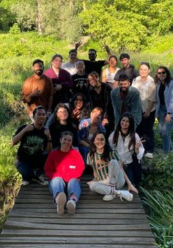 Grupa młodych ludzi stoi na drewnianym pomoście i pozuje do zdęcia. Wokół nich zielona łąka z wysokimi krzewami.