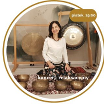 Kobieta w średnim wieku, z czarnymi włosami do ramion, ubrana w białą koszulkę z długim rękawem i spódnicę w kwieciste wzory. Kobieta siedzi na dywanach, w ręku trzyma kijek do uderzania w gonki i misy. Przed nią kilka mis, a za dnią dwa wiszące gongi.