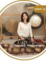 Kobieta w średnim wieku z czarnymi włosami. Ubrana w biały długi rękaw i wzorzyste spodnie. Siedzi na podłodze pokrytej dywanami. Za nią wiszą 2 gongi. Przed nią leży kilka mis.