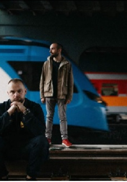 Mrokas stoi na peronie i Mrokas siedzi na peronie. W tle pociągi.