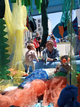 Fragment instalacji przypominającej dżunglę. W tle kilka zwiedzających osób, w tym małe dziecko bawi się wśród kolorowych tkanin.