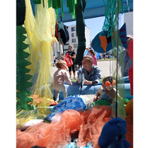 Fragment instalacji przypominającej dżunglę. W tle kilka zwiedzających osób, w tym małe dziecko bawi się wśród kolorowych tkanin.