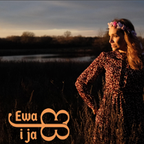 Kobieta w sukience w kwiaty i wiankiem na głowie patrzy w prawo z uśmiechem, w tle jezioro