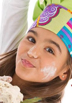 Dziewczynka ubrudzona mąką, trzyma surowe ciasto w ręce, na głowie ma kolorową czapkę kucharską. Obok rysunki czosnku, cytryny, liści, doniczki z ziołami i młynku do pieprzu.