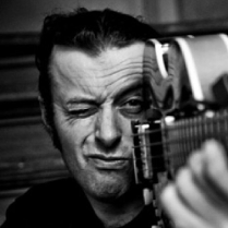 Tymon Tymański trzyma gitarę tak, jakby to była broń i celuje nią przed siebie mrużąc jedno oko