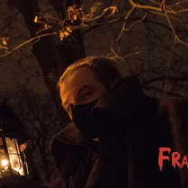 Mężczyzna w czarnym płaszczu, trzyma lampę naftową, za nim drzewo. Jest noc.