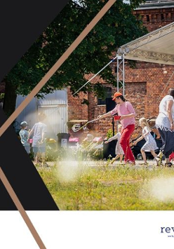 Zdjęcie buynku, a przed nim scena. Na niej śpiewają dzieci. Przed sceną rodzice z dziećmi bawią się bańkami mydlanymi.