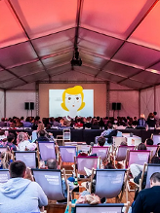 Ludzie siedzący na lrżakach i oglądający film w plenerze, pod namiotem.