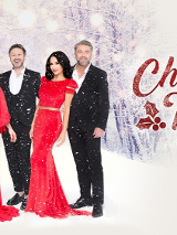 Trzech mężczyzn w garniturach oraz dwie kobiety w czerownych sukienkach pozują na tle śnieżnego lasu. Obok czerowne napisy.