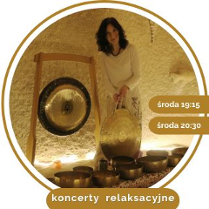 Kobieta w średnim wieku, w białym stroju, z czarnymi kręconymi włosami. Stoi przy rozłozonych złotych misach, trzyma duży gong w ręce. W tle wisi drugi gong.