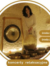 Kobieta w średnim wieku, w białym stroju, z czarnymi kręconymi włosami. Stoi przy rozłozonych złotych misach, trzyma duży gong w ręce. W tle wisi drugi gong.