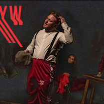 Zdjęcie przedstawia scenę teatralną w ciemnej tonacji. Na pierwszym planie stoi mężczyzna w białej koszuli z szelkami i czerwonych spodniach dresowych, trzymający w dłoni kaszkiet i drapiący się po głowie. W tle, w półmroku, widoczne są dwie inne postacie oraz fragment drewnianego stołu. W górnym lewym roku tytuł spektaklu zapisany czerwoną, niejako rozmytą czcionką.