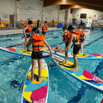 Uczestnicy zajęć stoją w kapokach na deskach SUP. Trener w wodzie.