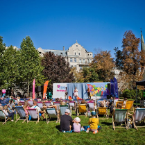 Ludzie siedzą na zielonej trawie w parku. Patrzą na scenę, na której występują ludzie. Scena jest ozdobiona logo, kolorowymi rysunkami.
