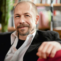 Profesor siedzi na kanapie, na tle regaów z książkami.