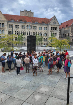 Grupa wycieczkowa stoi przy pomniku kryptologów. Pomnik składa się z prostokątnej bryły z wyrytymi cyframi. Wśród nich są nazwiska kryptologów. Wycieczkowicze są w różnym wieku, ale wspólnie słuchają przewodniczki. W tle budynek Banku Pocztowego na ul. Święty Marcin.