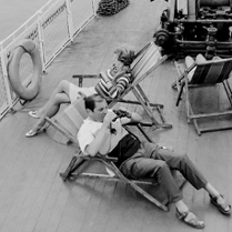 Kadr przedstawia dwie osoby, kobietę i mężczyznę, leżących na leżaczkach na pokładzie statku wycieczkowego. Patrzymy na nich z lotu ptaka.