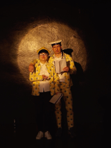 Zdjęcie z przedstawienia - kobieta i mężczyzna stoją obok siebie w zółtych strojach i czapkach na głowach, każde z nich trzyma w dłoni otwarty zeszyt.