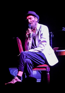 Zdjęcie ze spektaklu: przy pianinie, odwrócony do niego tyłem, na krześle siedzi mężczyzna z mikrofonem w ręce.