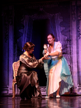 Zdjęcie ze spektaklu - dwie kobiety na scenie, jedna siedzi na krześle, druga stoi i trzyma w rękach lalkę.