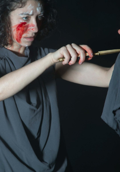 Zdjęcie ze spektaklu - kobieta z zakrwawioną twarzą w jednej ręce trzyma lalkę, a w drugiej sztylet, który przykłada do lalki.